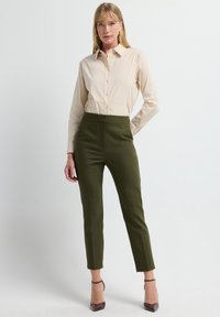 Hellbeige Hemdbluse mit langen Ärmeln, kombiniert mit taillierten, olivgrünen High-Waist-Hosen und braunen High Heels.