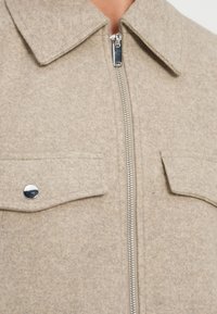 Μπεζ μπουφάν από μάλλινο μείγμα με γιακά, εμπρόσθιο φερμουάρ και δύο τσέπες με καπάκι, με επικάλυψη από ασημί υλικά και απαλή υφή.