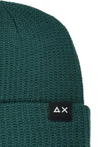 Berretto lavorato a maglia verde con risvolto, caratterizzato da un'etichetta rettangolare nera con icone geometriche bianche. La trama è morbida con una lavorazione leggermente allentata.