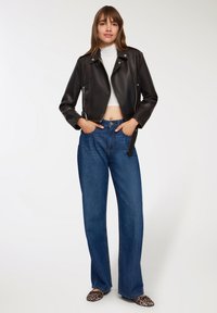 Kobieta w czarnej skórzanej kurtce, białym topie w stylu crop, niebieskich szerokich dżinsach i butach na leopardzi wzór, stoi z rękami w kieszeniach.