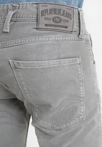 Vaqueros de mezclilla gris con bolsillos traseros y una etiqueta de marca de tela. El material tiene una ligera textura y la costura es visible a lo largo de las costuras.