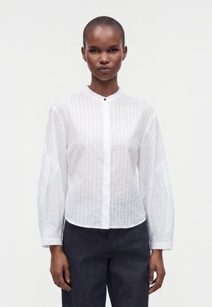 Femme aux cheveux courts portant un chemisier blanc à manches longues avec de fines rayures verticales et un pantalon foncé, debout devant un fond uni.