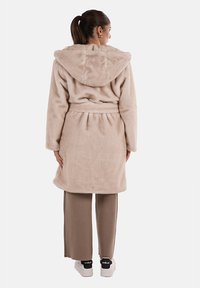 Accappatoio morbido beige, con cappuccio e una texture pelosa, in vita legato con cinghia e maniche lunghe; abbinato a pantaloni larghi color marrone chiaro e sneakers bianche.