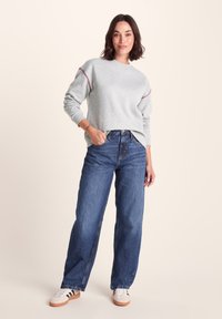 Gray sweatshirt with red stitching accents, paired with high-waisted blue denim jeans. Model stands in white sneakers with brown soles.