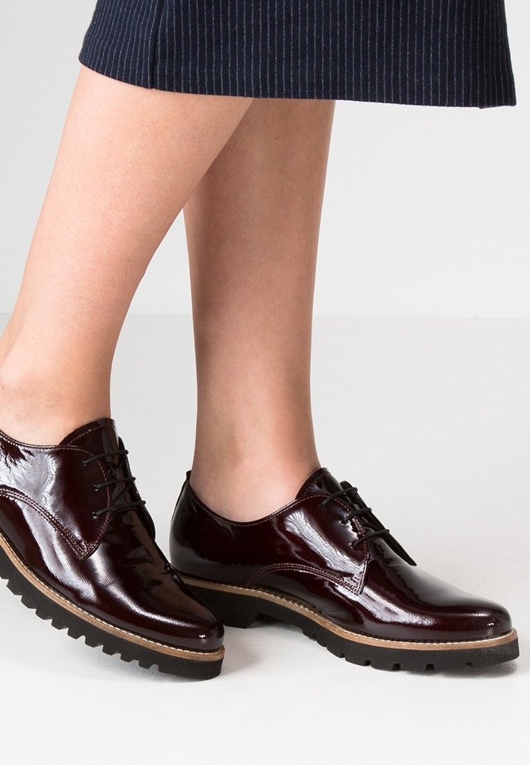 Chaussures Oxford en cuir verni bordeaux foncé à lacets avec semelles épaisses en caoutchouc noir, portées avec une jupe marine à fines rayures sur des jambes nues.