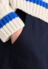 Griuvęs megztinis su mėlynomis juostelėmis ant rankovių, dėvimas virš tamsiai mėlynos medžiagos, su ranka, matoma šoninėje kišenėje.