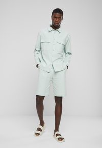 Lysegrønn skjorte med to brystlommer, kombinert med matchende lysegrønne shorts og beige sandaler. Glatt stoff og avslappet passform.