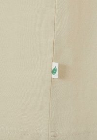 Beige pamut póló oldalsó címkével, amelyen zöld levél szimbólum található. Puha textúra, egyszerű design és látható varrási részletek.