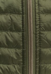 Primo piano di una giacca imbottita verde oliva, con un tessuto testurizzato, un motivo a cuciture orizzontali e una cerniera metallica al centro.