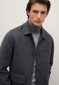 Grauer Wollmantel mit Kragen, vorderer Knopfleiste und zwei aufgesetzten Taschen. Getragen über einem hellgrauen, gerippten Rollkragenpullover.