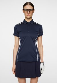 Mörkblå polo med vita detaljer, korta ärmar och fyra knappar. I kombination med en plisserad mörkblå kjol. Vit handske på vänster hand.