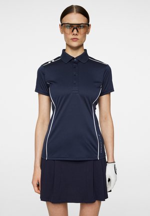 Polo shirt blu navy con dettagli bianchi, maniche corte e quattro bottoni. Abbinato a una gonna blu navy a pieghe. Guanto bianco sulla mano sinistra.