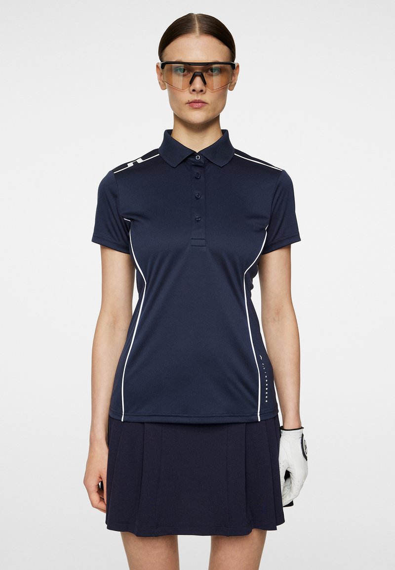 Mörkblå polo med vita detaljer, korta ärmar och fyra knappar. I kombination med en plisserad mörkblå kjol. Vit handske på vänster hand.