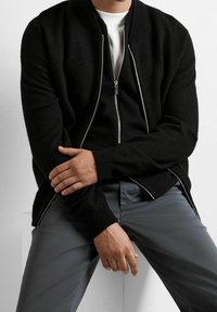 Svart bomberjacka i texturerat tyg med dragkedjeslutning, ribbad krage och kontrasterande silverdetaljer. Bärs över en vit skjorta.