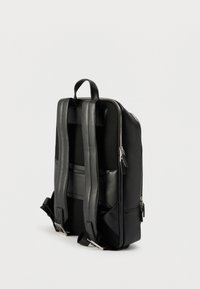 Sac à dos en cuir noir avec bretelles rembourrées, panneau arrière en mesh, deux fermetures éclair argentées et une poignée de transport supérieure, incliné sur un fond neutre.