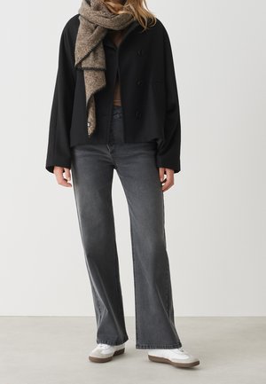 Osoba w czarnej, zapinanej na guziki kurtce, beżowym szalu, szarych dżinsach o szerokich nogawkach i białych trampkach na jednolitym tle.