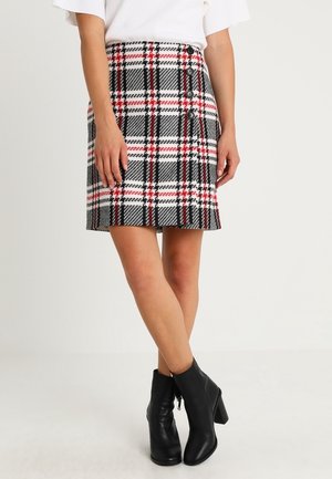 Mujer que lleva una falda de cuadros negra, blanca y roja de talle alto con botones en el costado, combinada con botas negras hasta el tobillo y una camiseta blanca.