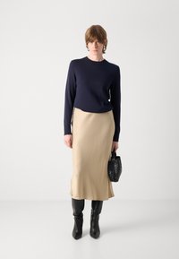 Marineblå strikkegenser, beige satengskjørt, svarte knelange støvler og en teksturert svart håndveske. Minimalistisk design med glatt stoff og elegant silhuett.