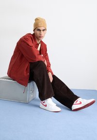 Il modello indossa una giacca leggera rossa, pantaloni in corduroy marroni e sneakers bianche con accenti rossi. Un beanie beige completa l'outfit.