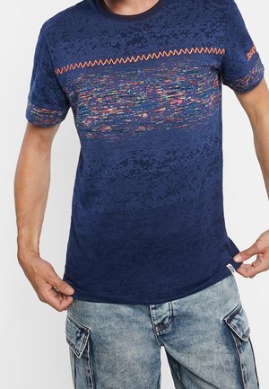 Mężczyzna w granatowym T-shircie z wzorem w wielobarwne pikselowe motywy i pomarańczowym zygzakowatym detalem, w połączeniu z jasnoniebieskimi jeansami typu cargo.