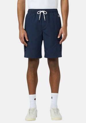 Uomo che indossa pantaloncini cargo blu navy con coulisse, calzini bianchi con logo, sneakers bianche e una maglietta bianca infilata dentro.