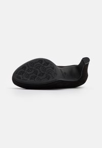 Zapato mocasín de ante negro con un diseño curvado, que presenta una suela de goma con patrón y un pequeño tacón en la parte trasera para mayor soporte.