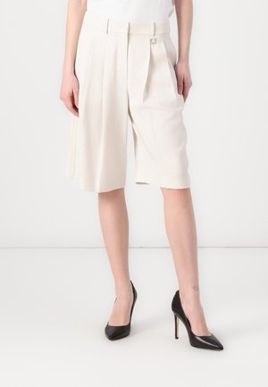 Beige tailored shorts with pleats, wide leg, and a belt detail. Paired with black high-heeled shoes. Smooth, lightweight fabric.