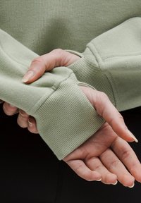 Svetlá zelená mikina s rebrovanými manžetami, textúrovanou tkaninou a voľným strihom. Ruky sú vidieť, ako držia látku na rukáve.