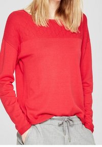 Pull en maille rouge avec une partie supérieure texturée et des manches longues, associé à un pantalon gris à cordon de serrage. Le tissu semble doux et léger.