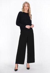 Marineblauwe gebreide cardigan met knopen, gecombineerd met zwarte rechte broek en zwarte hoge hakken. Model staat tegen een witte achtergrond.