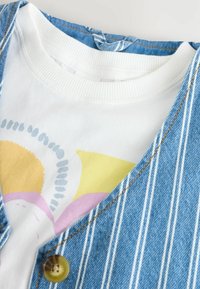 Denim prsluk s bijelom majicom s grafikom ispod. Prsluk ima plave i bijele vertikalne pruge, s narančastim šavovima i smešnim dugmetom.