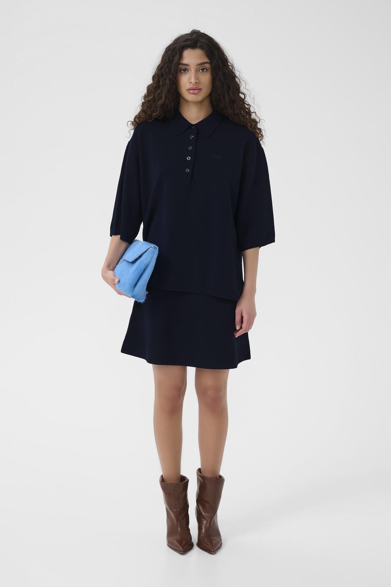 Polo bleu marine à trois boutons, assorti à une jupe coordonnée. Le mannequin tient une pochette bleu clair et porte des bottines marron.