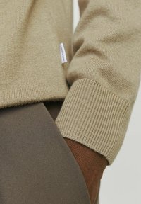 Beige Strickpullover mit gerippten Bündchen, kombiniert mit einem braunen Stoff darunter. Auffällige Textur in beiden Materialien; kleines Markenetikett sichtbar.