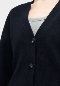 Cardigan en maille bleu marine avec deux boutons noirs sur un haut côtelé gris clair à col rond, montré de la poitrine jusqu'au cou.