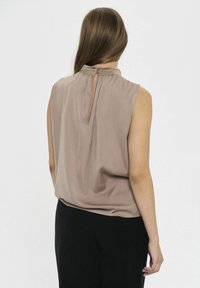 Blusa sin mangas color beige con cuello alto y abertura en forma de ojo de cerradura en la parte trasera, hecha de una tela suave y ligera que cae con delicadeza.