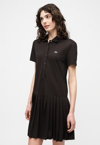 Jeune femme aux cheveux bouclés portant une robe polo noire à manches courtes avec une jupe plissée et un petit logo sur la poitrine, se tenant légèrement de profil.