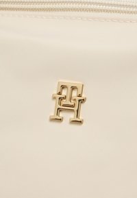 Υφάσμα σε κρεμ χρώμα με λεία υφή, στο οποίο υπάρχει χρυσό μεταλλικό λογότυπο με σχήμα αλληλοσυνδεόμενων γραμμάτων "H" και "H".