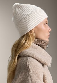 Berretto bianco a coste con punta, indossato con una giacca di pile beige a collo alto con superficie testurizzata e zip argentata.