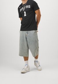 Schwarzes T-Shirt mit "SPURS" und der Nummer 1 in Weiß. Helle Denim-Shorts, knielang. Weiße Sneakers mit blauen Akzenten. Einfaches Perlenhalsband.