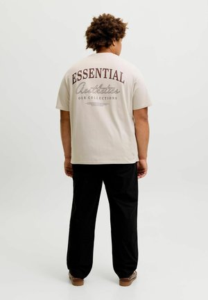 Person mit lockigem Haar steht mit dem Rücken zur Kamera, trägt ein beiges T-Shirt mit dem Schriftzug „ESSENTIAL Aesthetics Our Collections“, schwarze Hose und braune Schuhe.
