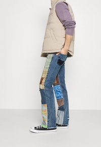 Vaqueros de denim con un diseño de patchwork que presenta múltiples colores y patrones. Combinados con un chaleco acolchado en un beige claro y zapatillas negras.