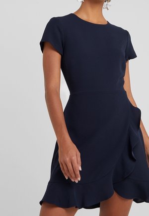 Kobieta w granatowej sukience z krótkim rękawem, okrągłym dekoltem i warstwowym, falbankowym dołem, stojąca na jednolitym jasnym tle.