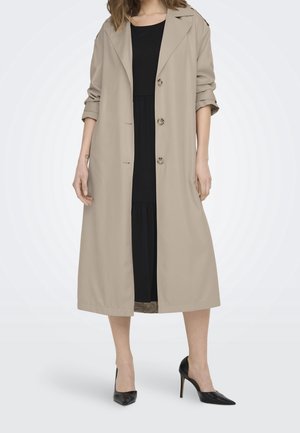 Femme portant un long trench-coat beige par-dessus une robe noire et des chaussures à talons hauts noires, debout devant un fond blanc.