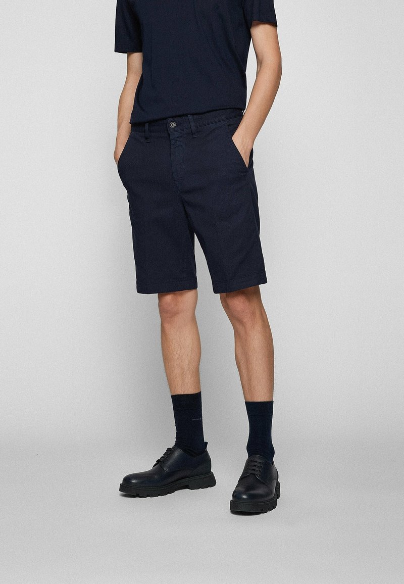 Personne portant un short noir, des chaussettes noires et des chaussures noires lacées, debout les mains dans les poches devant un fond clair uni.