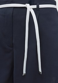 Cordon blanc noué en un nœud lâche sur un pantalon en tissu bleu marine texturé avec passant de ceinture visible et poche latérale.