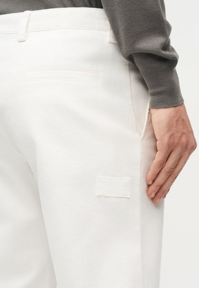 Gros plan sur le bas du torse d'une personne portant un pantalon écru avec une poche arrière et une chemise grise à manches longues, la main reposant près de la cuisse.
