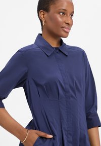 Granatowa sukienka koszulowa z kołnierzykiem i krótkimi rękawami, z dopasowanym gorsetem i marszczoną spódnicą. Gładka faktura, posiada boczne kieszenie.