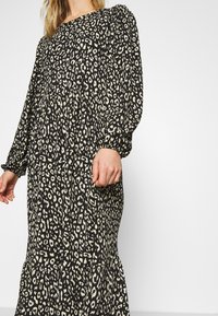 Femme portant une robe à manches longues imprimé léopard noir et blanc avec des poignets froncés et une jupe à volants, sur un fond blanc uni.