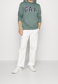 Felpa con cappuccio verde-grigio con logo "GAP", pantaloni bianchi larghi e sneakers bianche. La felpa presenta una tasca a marsupio e un cappuccio regolabile.