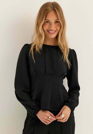 Donna sorridente con lunghi capelli biondi che indossa una camicetta nera a maniche lunghe con spalle arricciate, in piedi davanti a uno sfondo chiaro e uniforme.